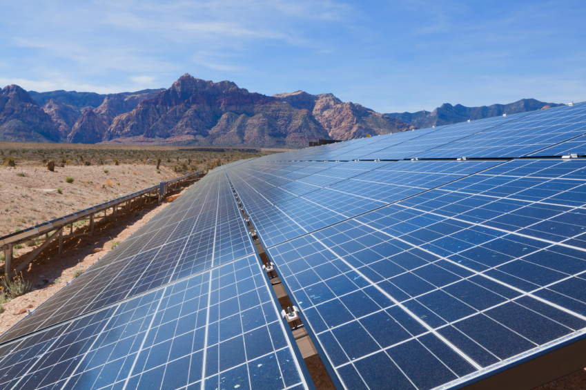 A large array of solar panels in the desert, with mountains visible in the background under a clear blue and partly cloudy sky.