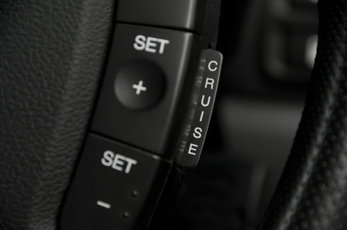 Close-up of cruise control buttons on a car steering wheel. The steering wheel has a textured, black leather surface. The buttons are marked with "SET", "+", "-", and "CRUISE".