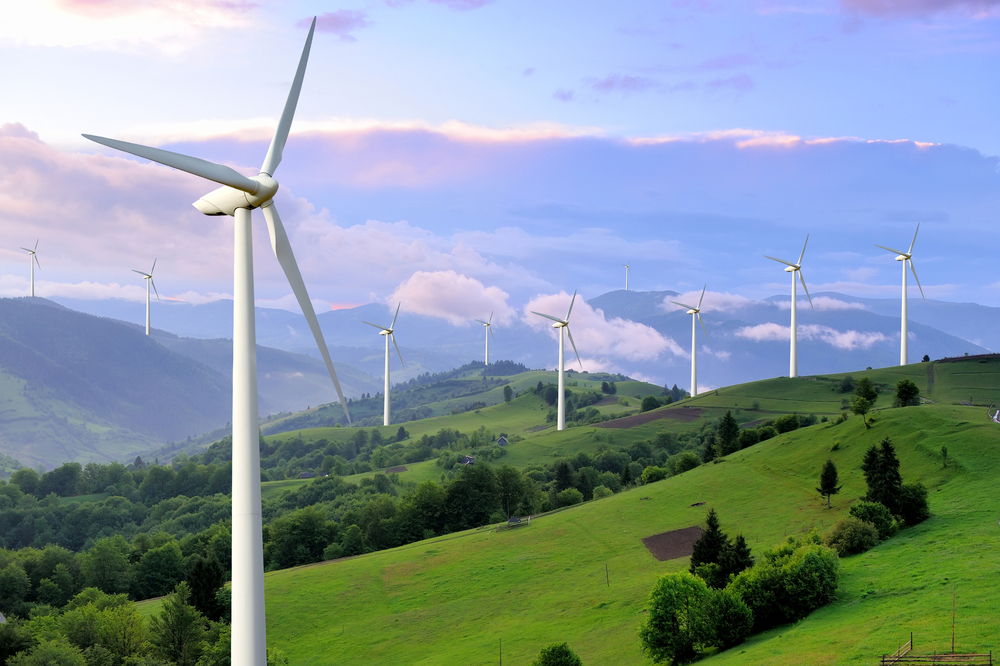 A landscape of green hills and trees with several wind turbines. One turbine is in the foreground, with others in the distance under a partly cloudy sky.